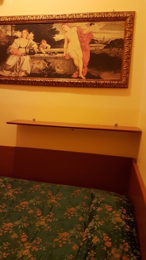 A room with a bed, a painting on the wall, and a shelf above the bed.