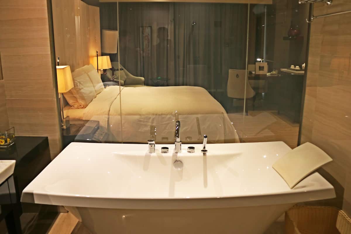 Ein modernes Badezimmer mit einer großen Badewanne, einer in Glas eingefassten Duschkabine und einem sichtbaren Schlafzimmer mit Bett und Nachttisch.