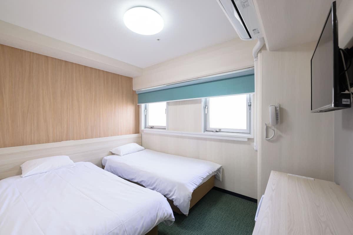A hotel room with two single beds, a window with a view, a flat-screen TV mounted on the wall, and a ceiling light.