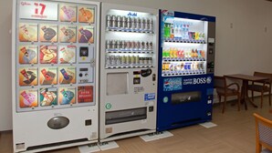 Máquinas de vendas com diversas bebidas e snacks, uma mesa de madeira e cadeiras em um quarto.