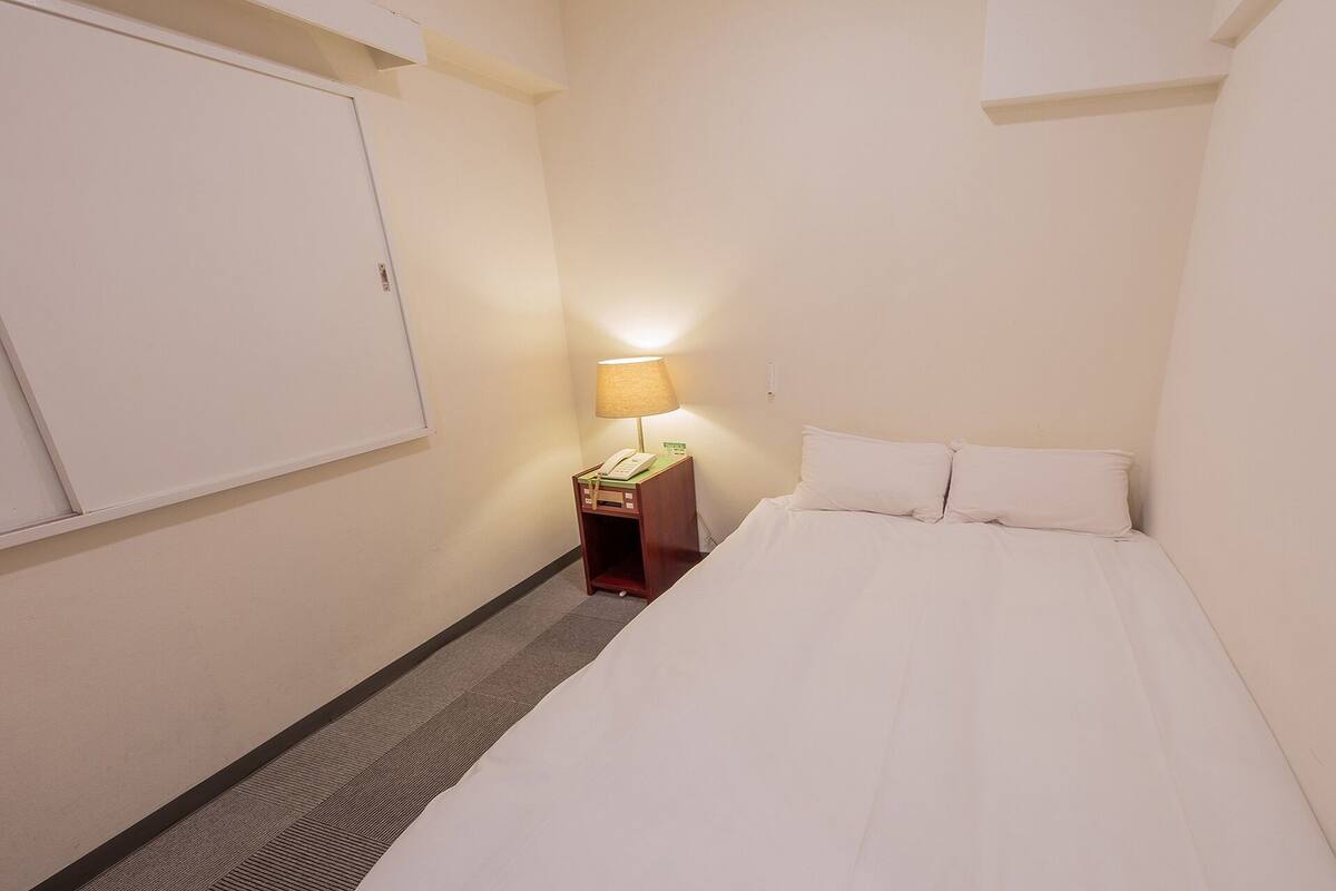 A small, narrow hotel room with a single bed, a bedside table with a lamp, and a wall-mounted phone.