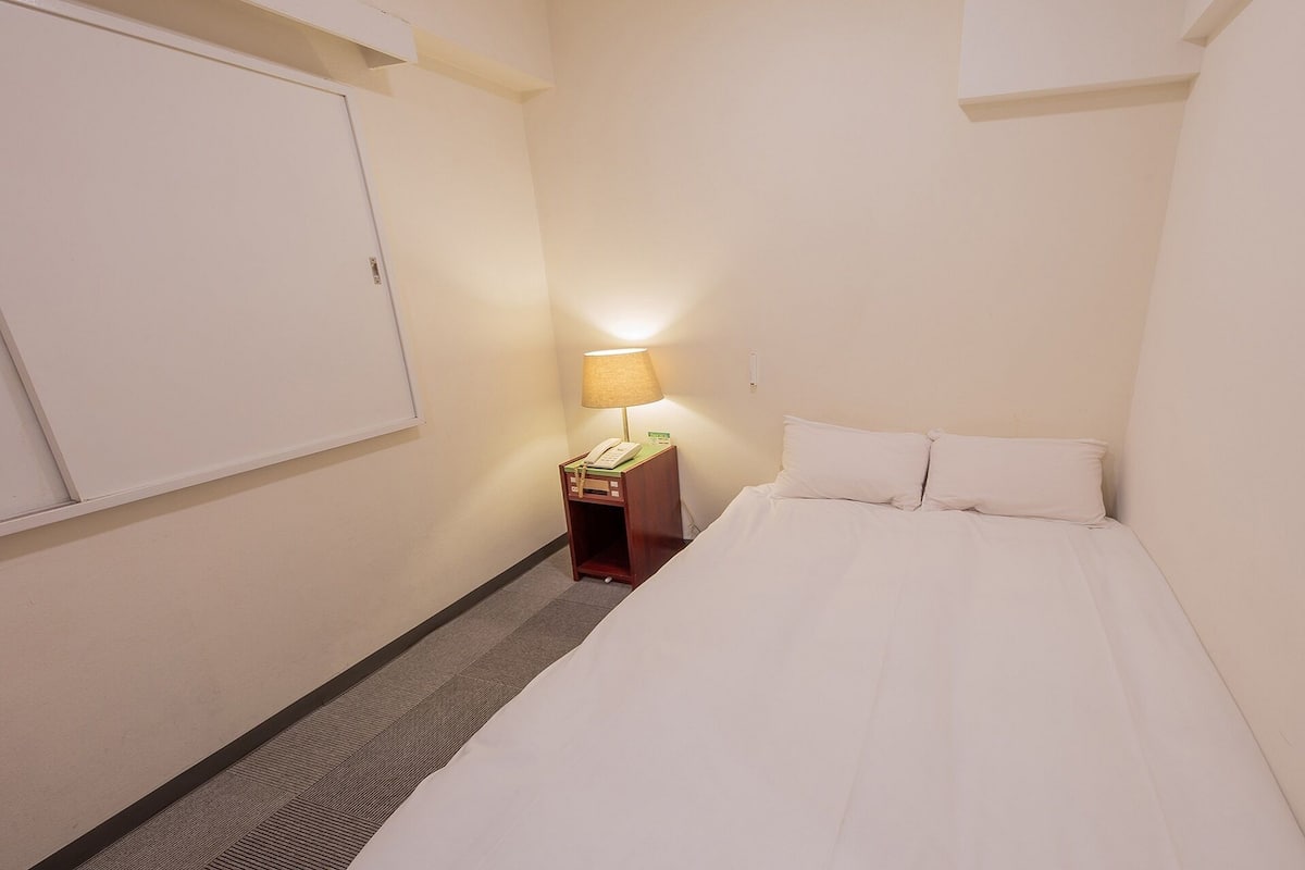 Ein kleines, schmales Hotelzimmer mit einem Einzelbett, einem Nachttisch mit Lampe und einem an der Wand befestigten Telefon.
