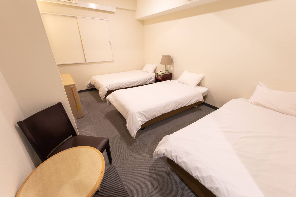 A hotel room with three single beds, a small table, a chair, and a bedside table with a lamp.