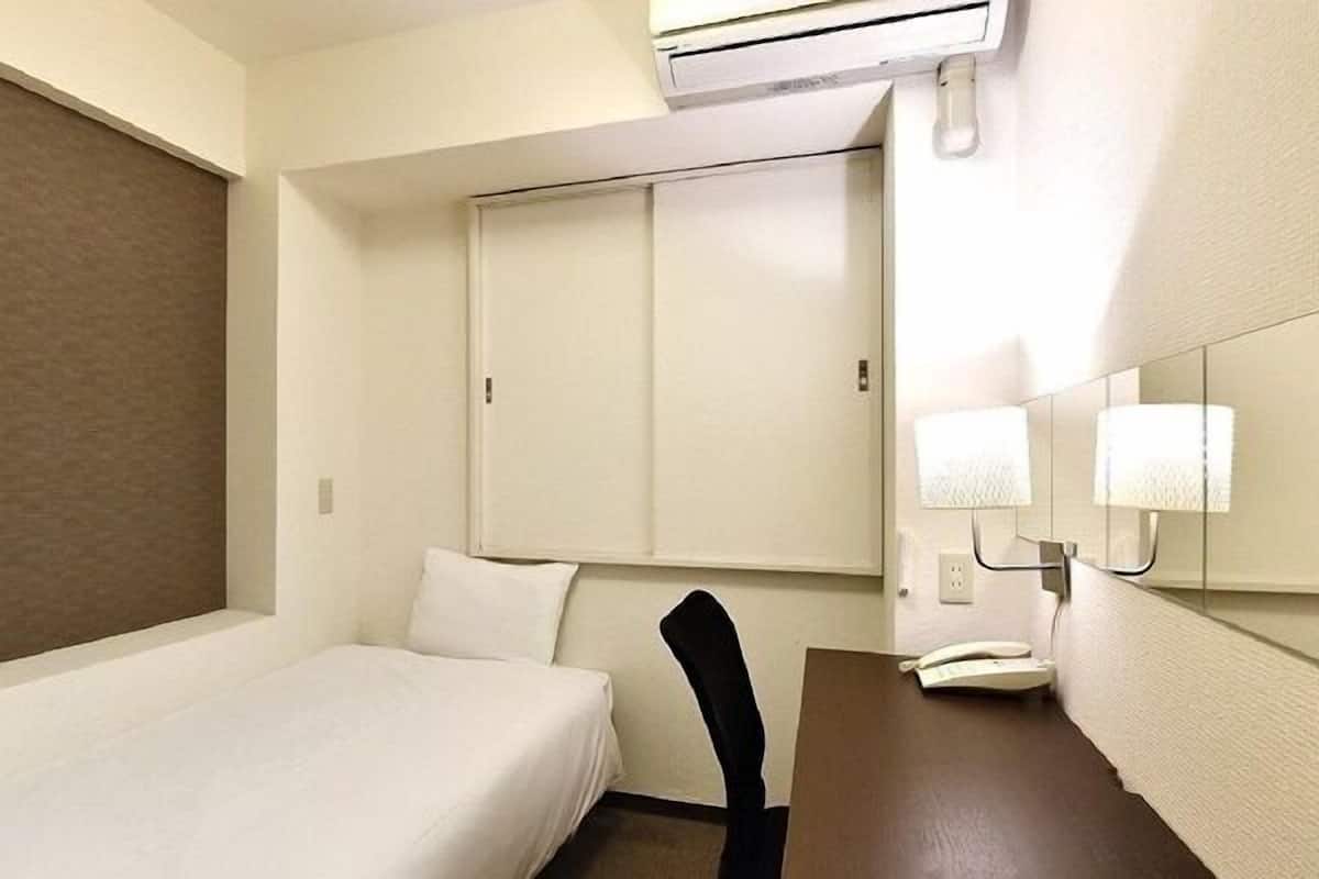 A hotel room with a single bed, a desk with a telephone, a chair, and a window with blinds.