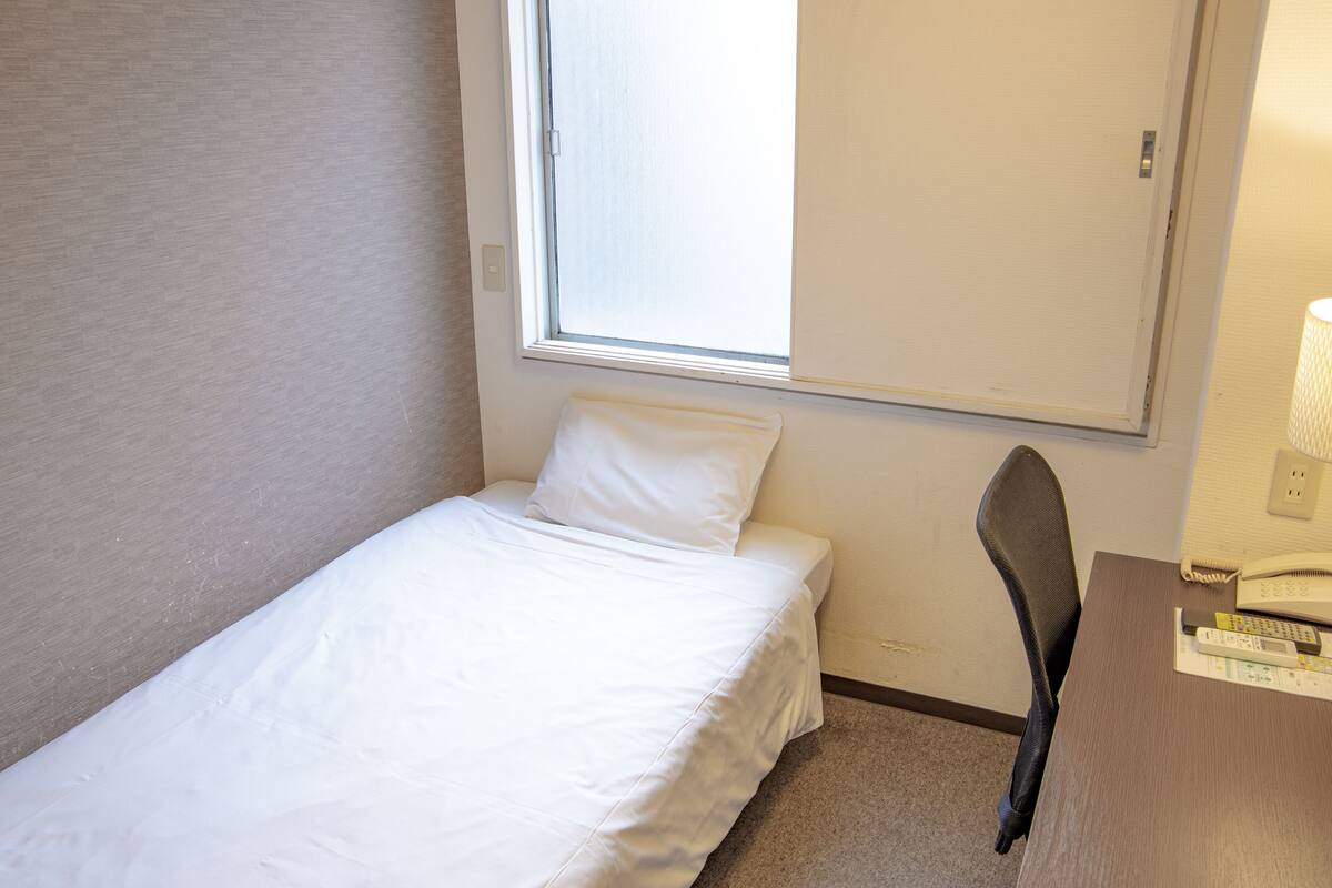 A small, single-bed room with a window, a desk with a lamp, and a chair.