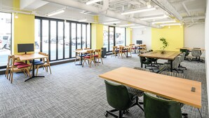 Um espaço de escritório moderno, com várias mesas e cadeiras, janelas amplas e uma parede de destaque em amarelo vibrante.