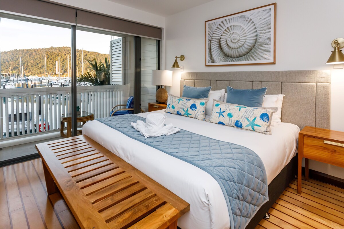 Een hotelkamer met een groot bed, een houten bankje, uitzicht op een jachthaven en een kunstwerk in een lijst aan de muur.