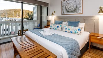 Habitación de hotel con una cama grande, un banco de madera, vista a un puerto deportivo y una obra de arte enmarcada en la pared.