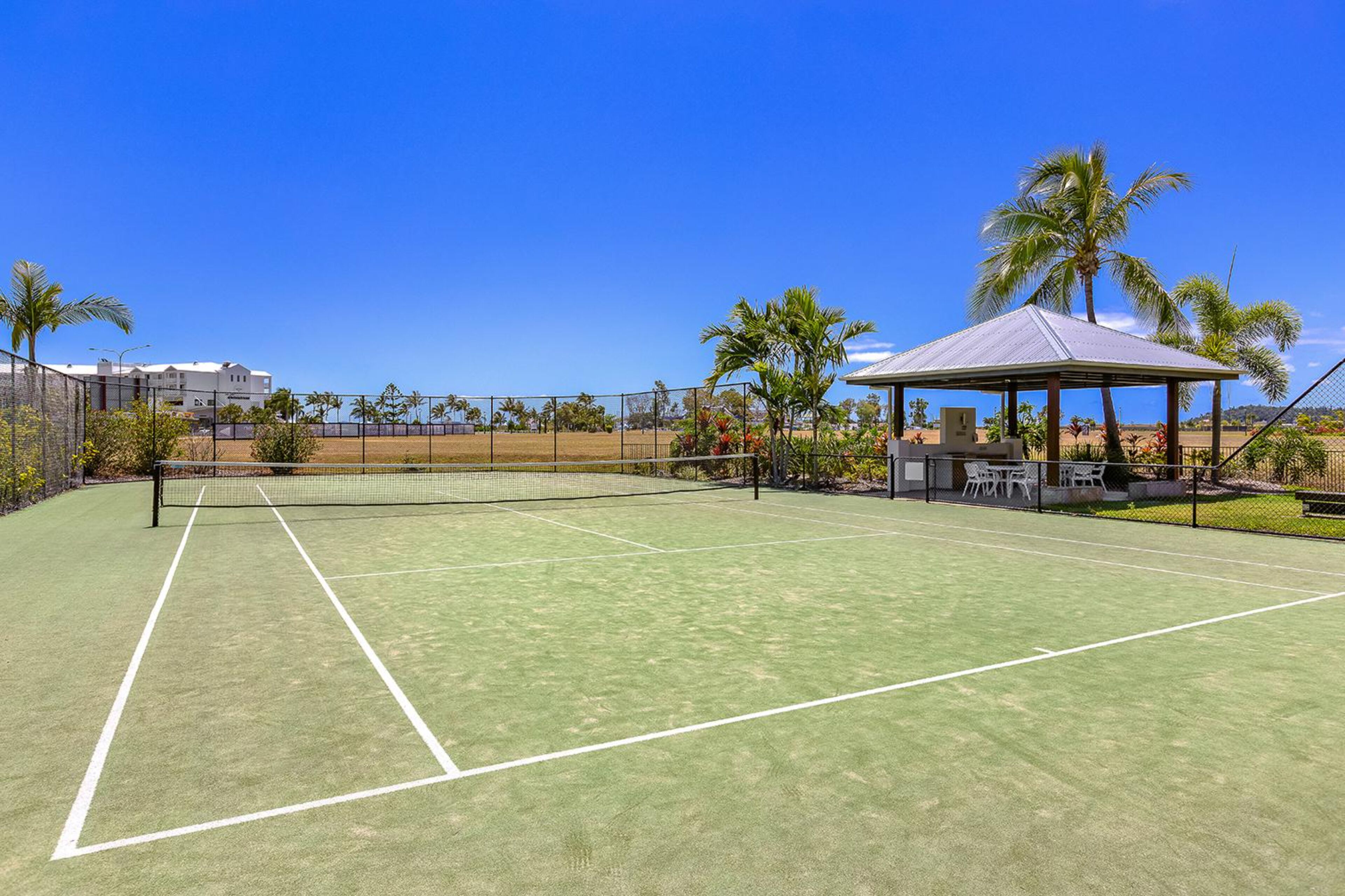 Una cancha de tenis con pabellón, palmeras y un edificio al fondo.
