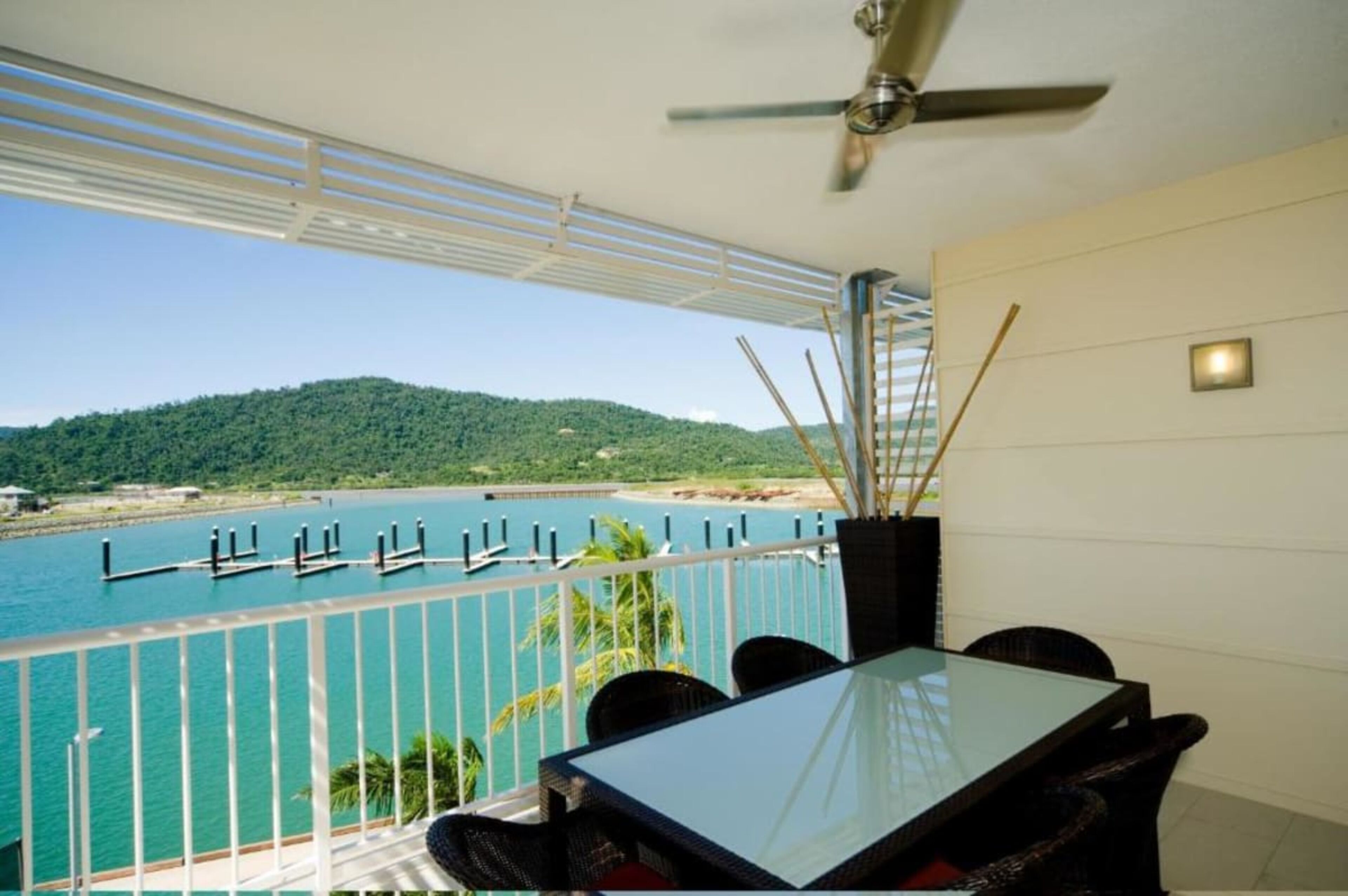A balcony with a glass table and chairs, overlooking a body of water with docks and a forested hill in the background.
