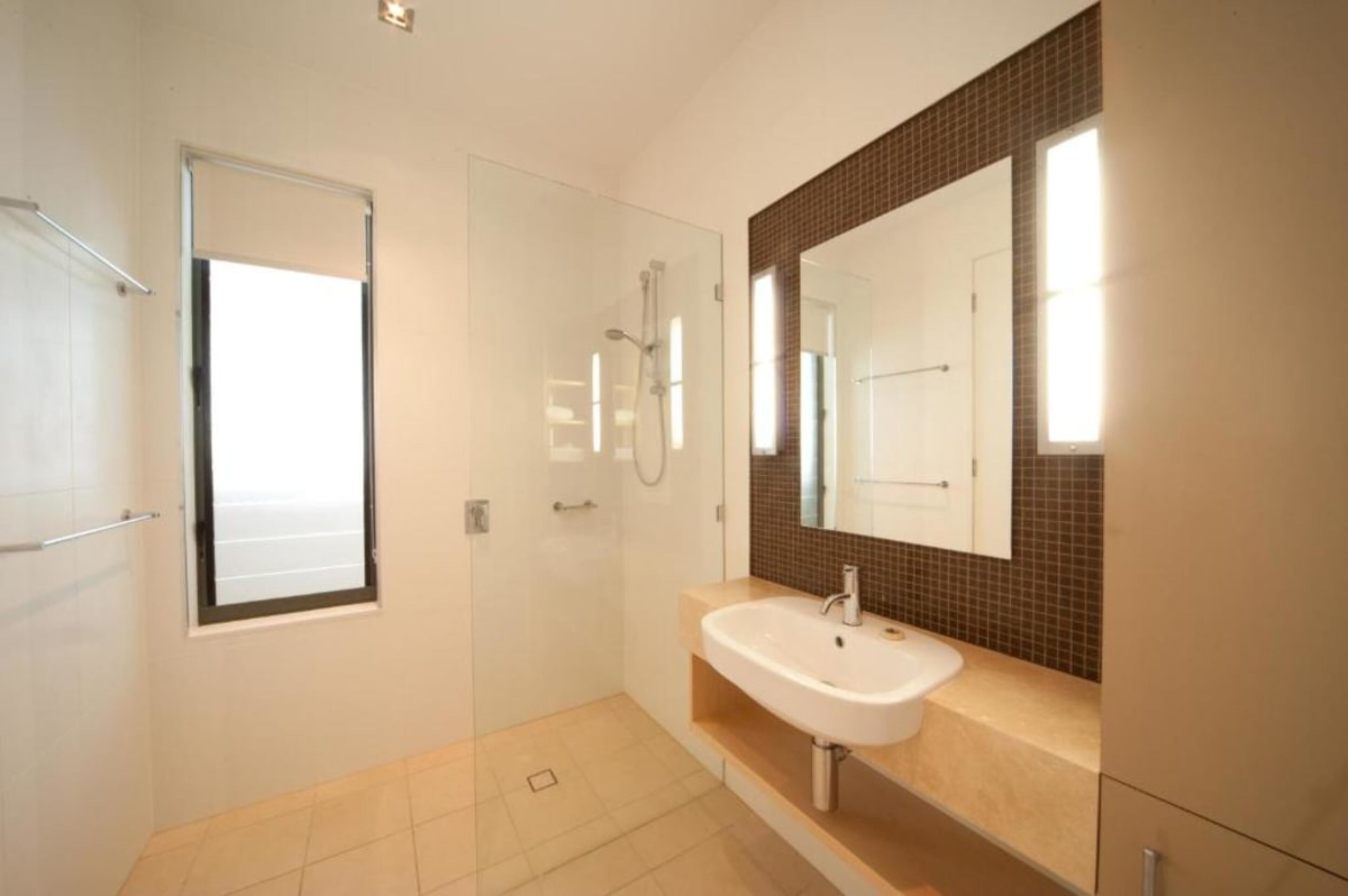 Un baño moderno con cabina de ducha de vidrio, un lavabo blanco y un espejo grande con salpicadero de baldosas marrones.
