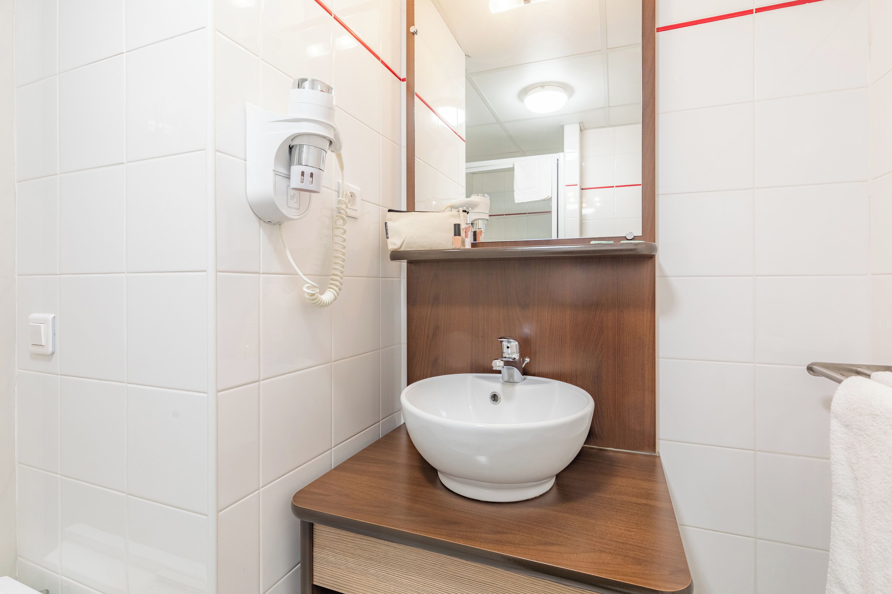 A bathroom with a white sink, wooden countertop, and a mirror above it. There is a hairdryer on the wall and a towel rack to the right.