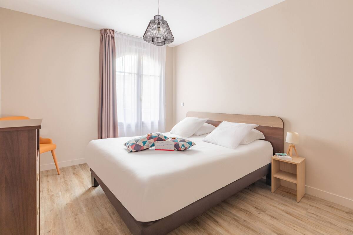 A neatly made bed with white linens and colorful pillows, a wooden nightstand with a lamp, and a desk with a chair.