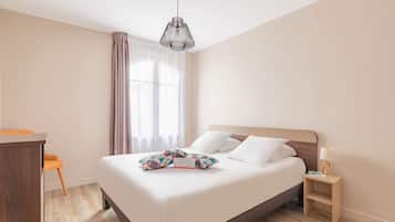 A neatly made bed with white linens and colorful pillows, a wooden nightstand with a lamp, and a desk with a chair.