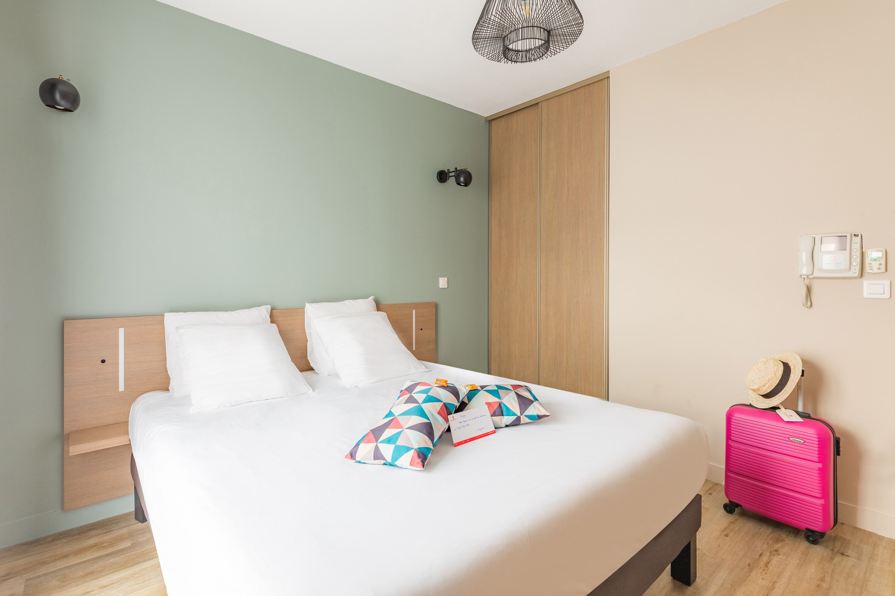A neatly made bed with white bedding and colorful geometric pillows, a wooden headboard, a wooden wardrobe, a pink suitcase, and a wall-mounted phone.