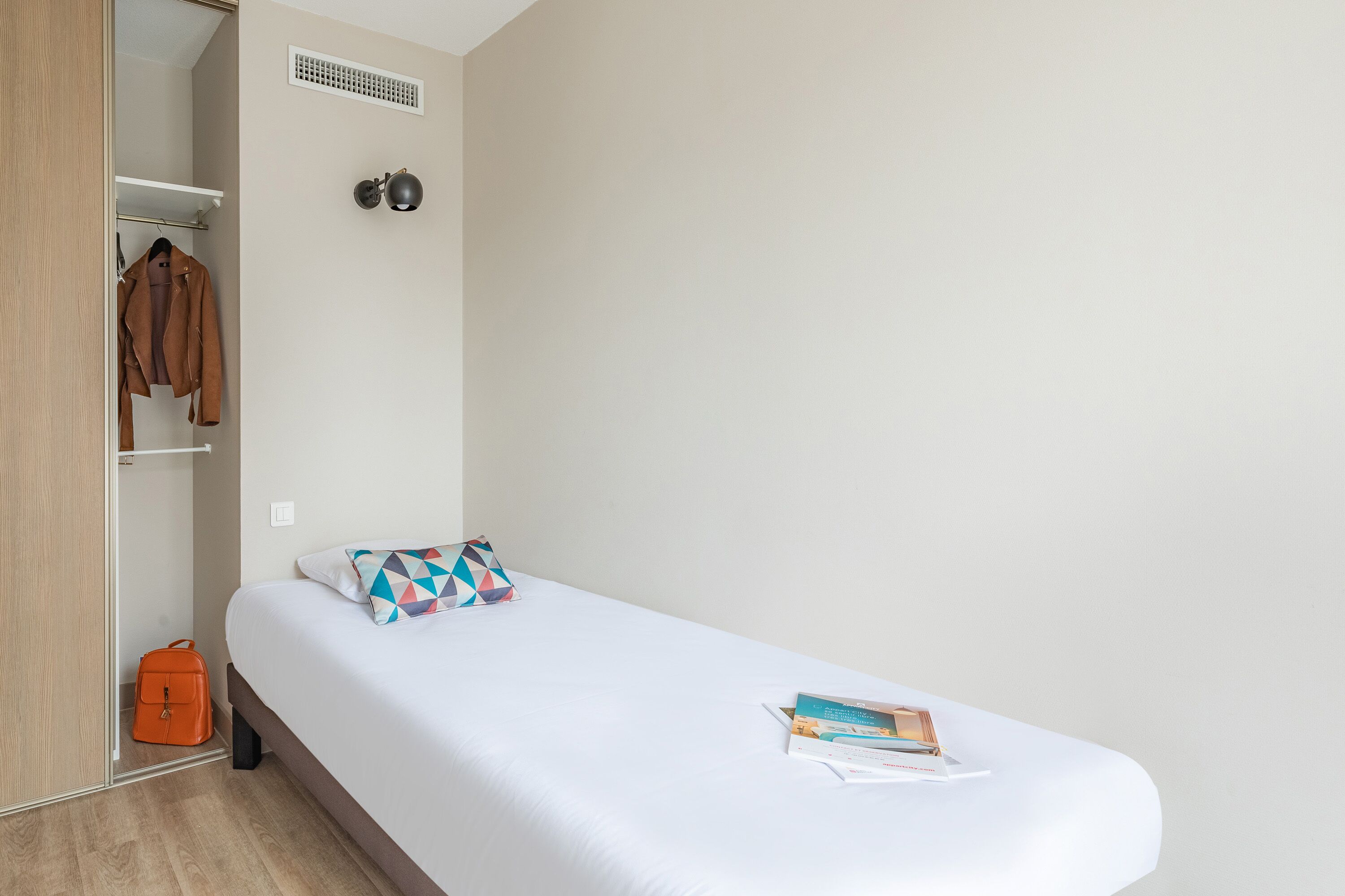 Habitación con cama individual, colchón blanco, almohada estampada con motivos geométricos, una pequeña bolsa naranja y un libro sobre la cama.