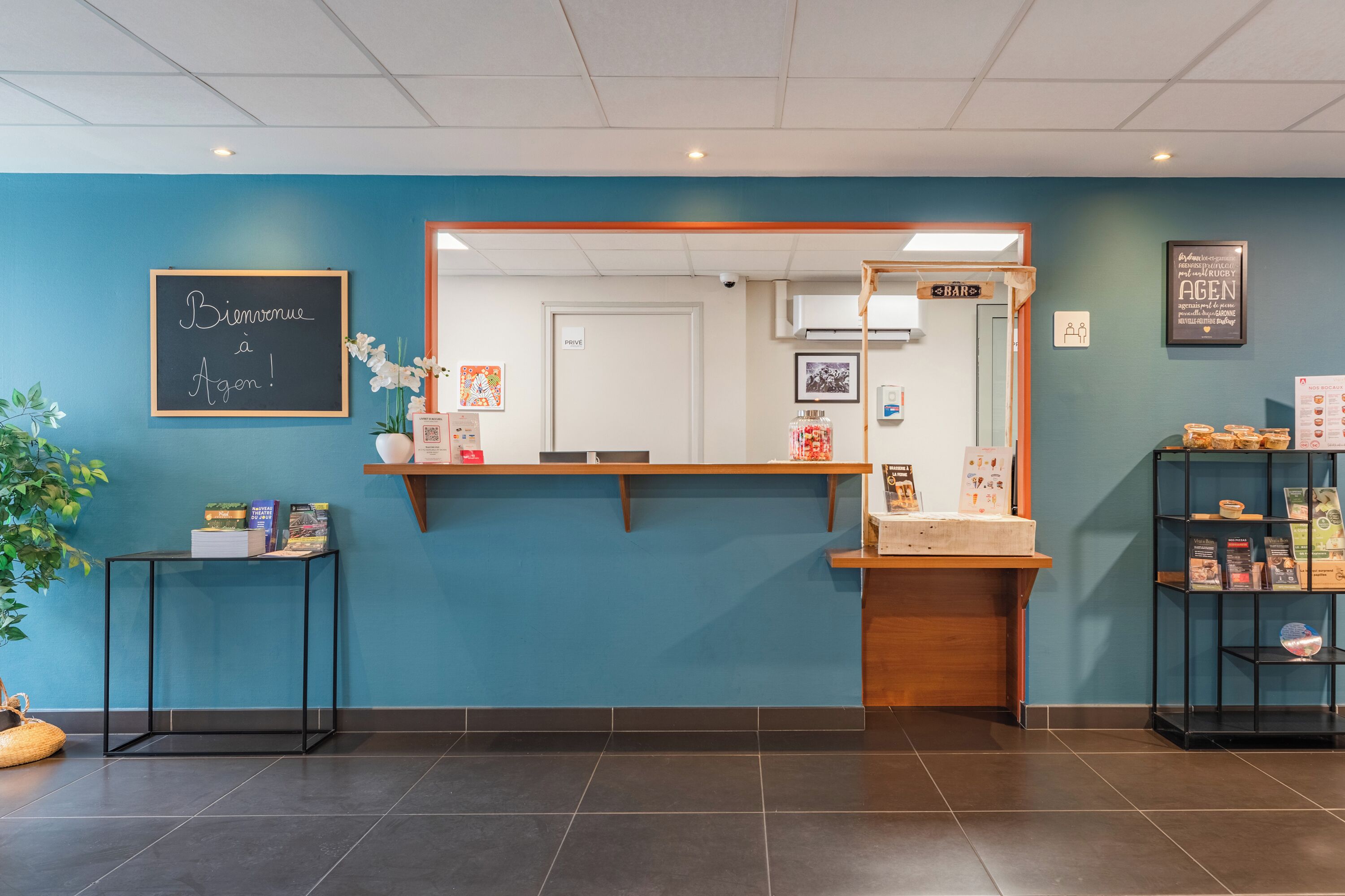 A reception area with a blue wall, a chalkboard with 'Bienvenue à Agen!', a wooden reception desk, and a shelf with books and a plant.