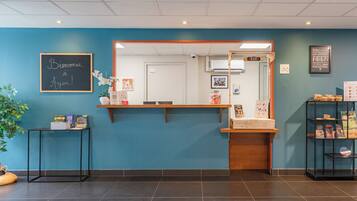 A reception area with a blue wall, a chalkboard with 'Bienvenue à Agen!', a wooden reception desk, and a shelf with books and a plant.
