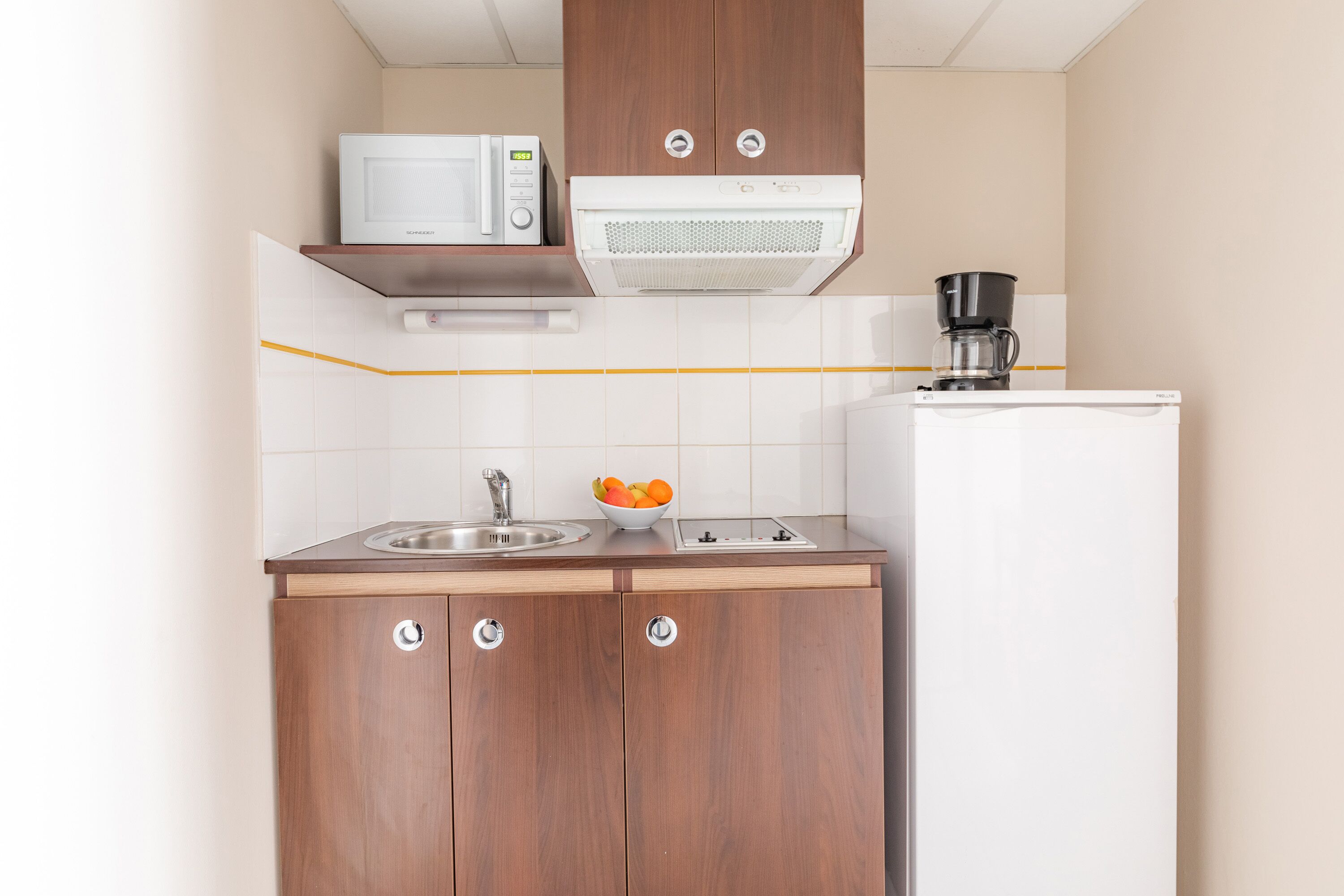 Een compacte keuken met een magnetron, koffiezetapparaat en een fruitschaal op het aanrecht.