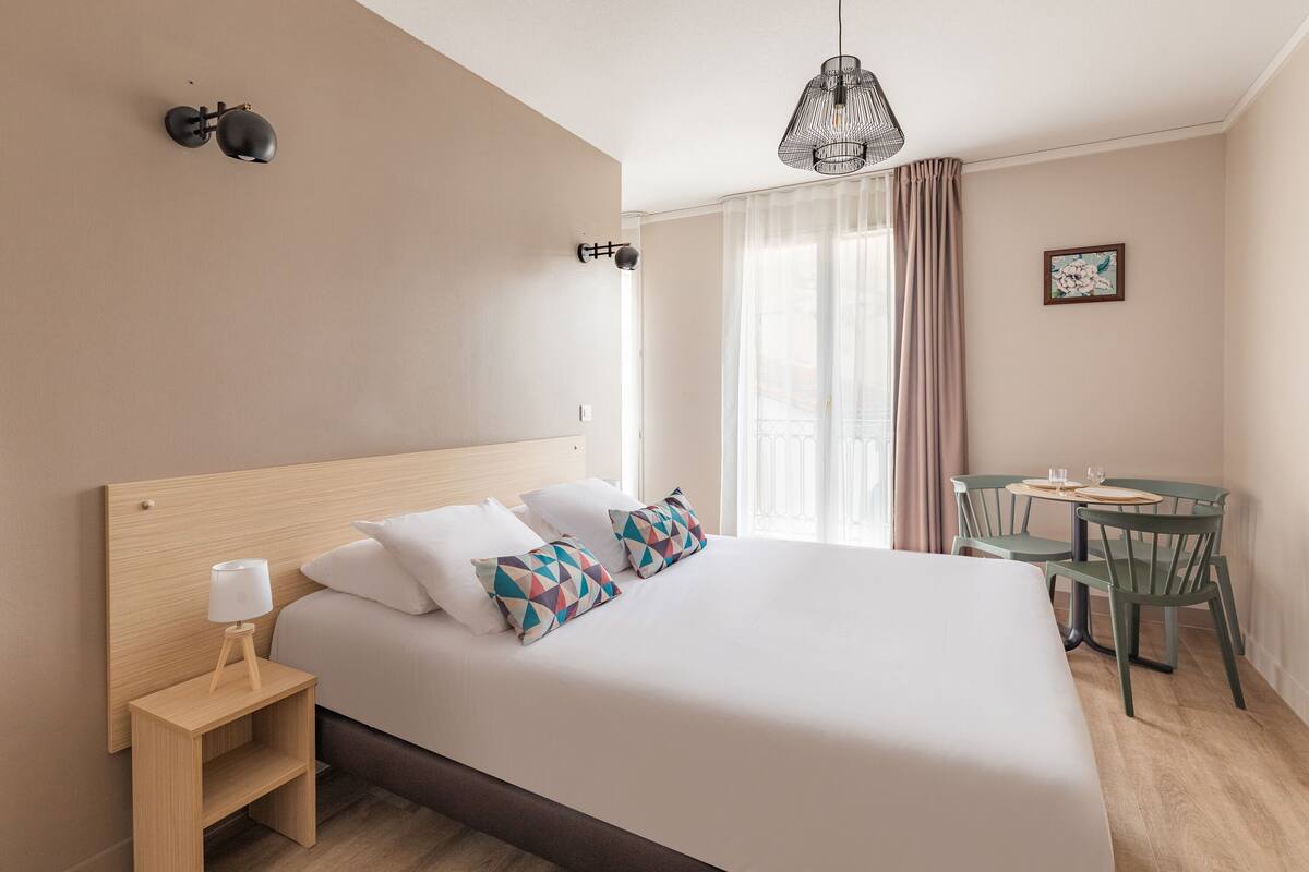 A neatly arranged hotel room with a large bed, bedside table, lamp, and a small table with chairs.
