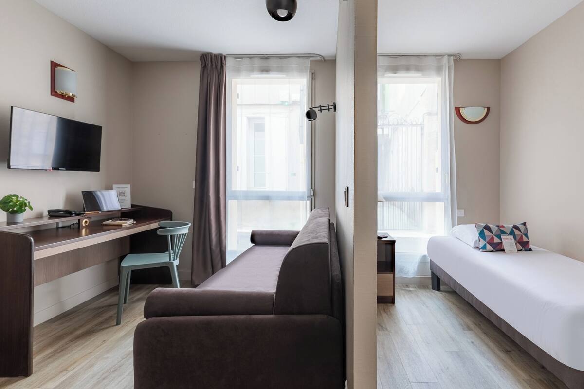 A modern hotel room with a sofa, a bed, a desk with a computer, a TV, and a window with curtains.