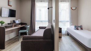 Una habitación de hotel moderna con sofá, cama, escritorio con computadora, televisor y ventana con cortinas.