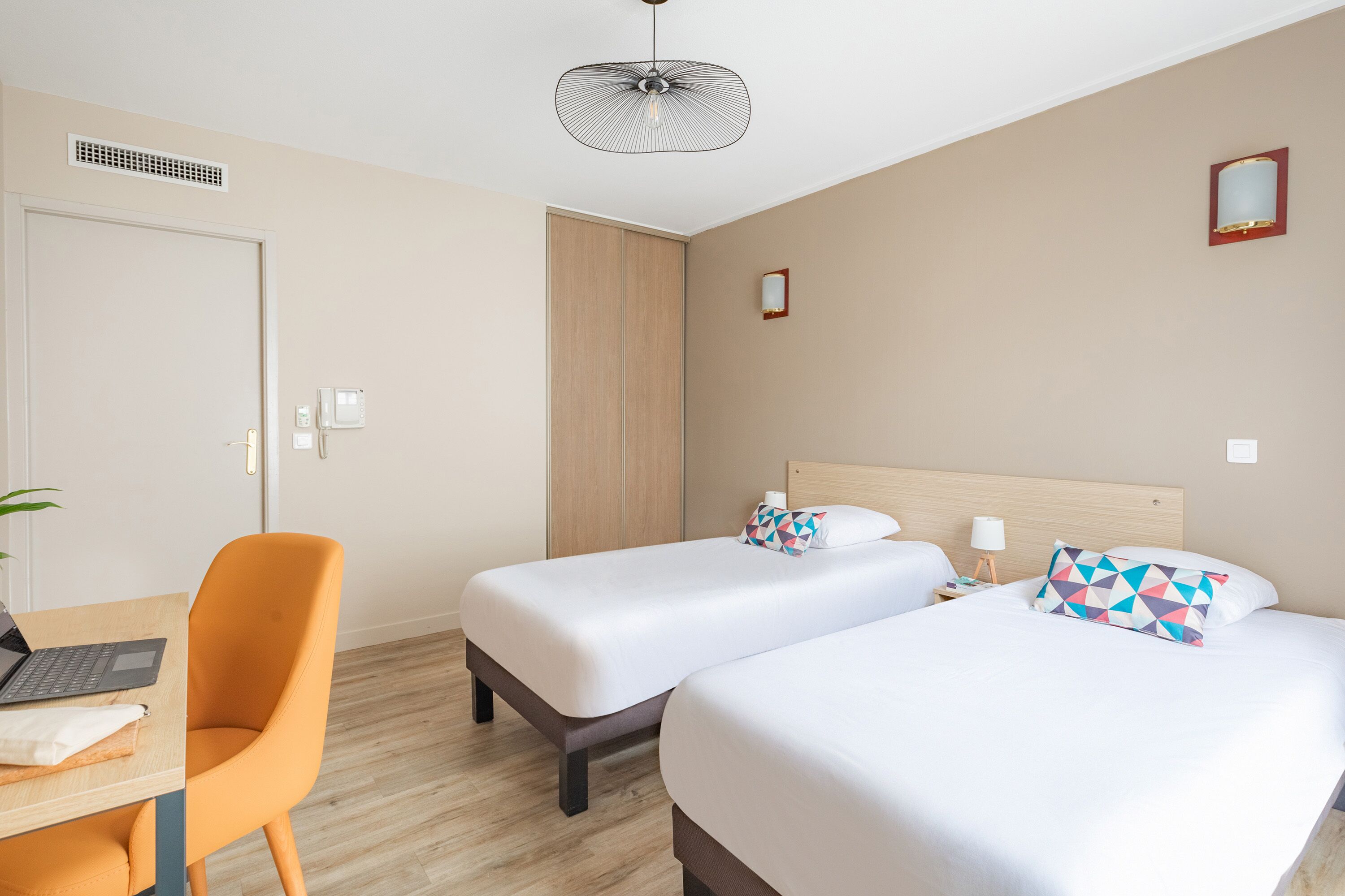 A hotel room with two beds, a desk with a laptop, an orange chair, a ceiling fan, and a small bedside table with a lamp.