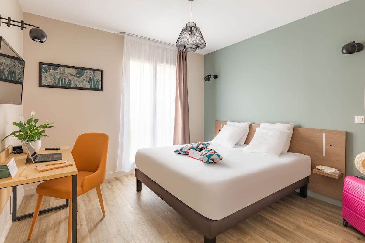 A neatly arranged hotel room with a bed, a desk with a laptop, a chair, a suitcase, and a plant.