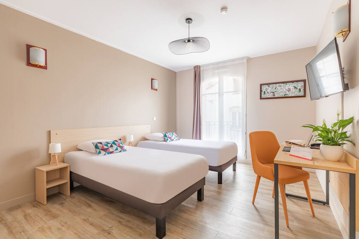 A hotel room with two beds, a desk with a chair, a TV, and a window with curtains.
