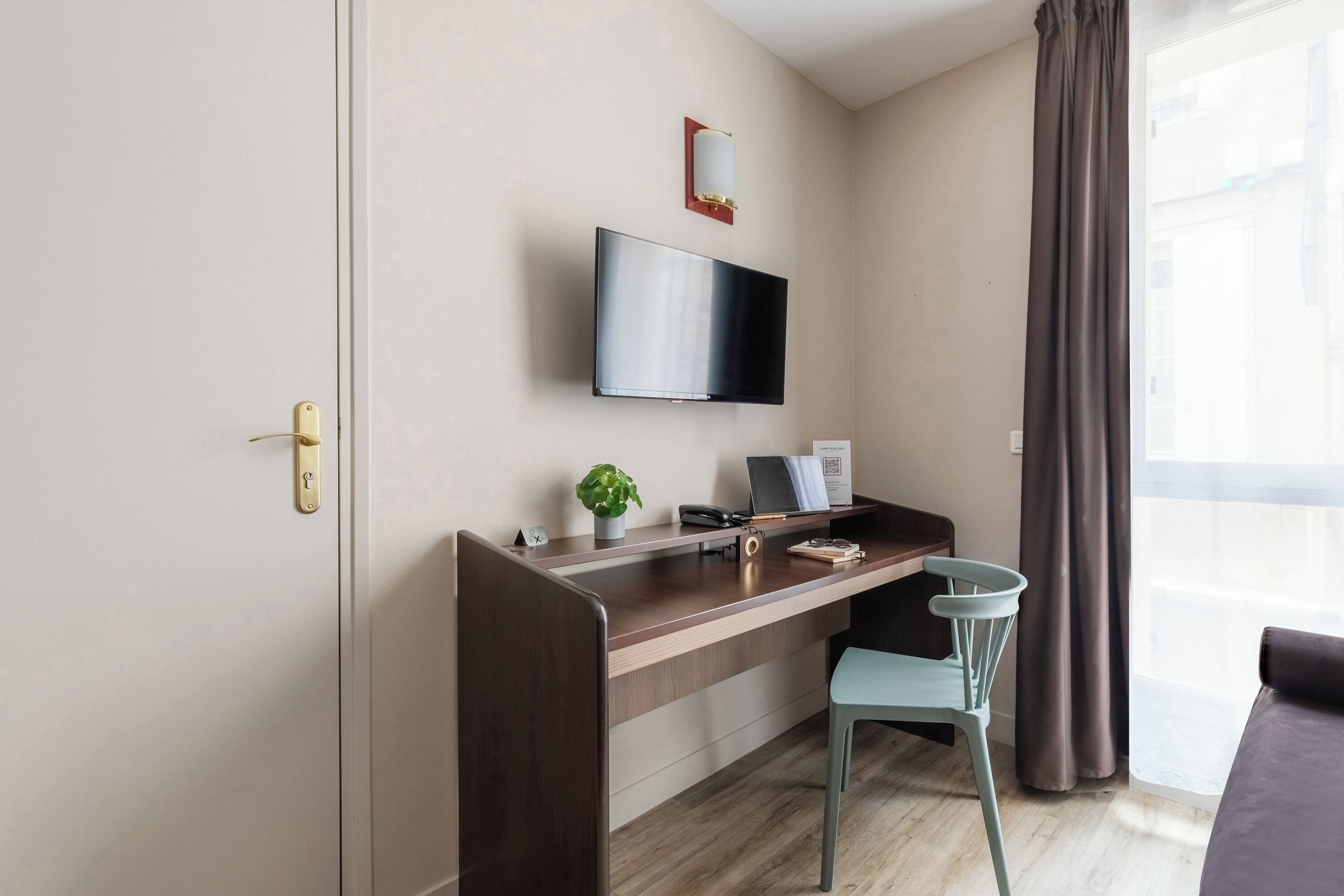 Una habitación moderna con un escritorio de madera, un televisor montado en la pared, una pequeña planta y una ventana con cortinas.