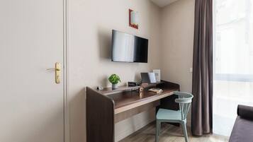 Una habitación moderna con un escritorio de madera, un televisor montado en la pared, una pequeña planta y una ventana con cortinas.