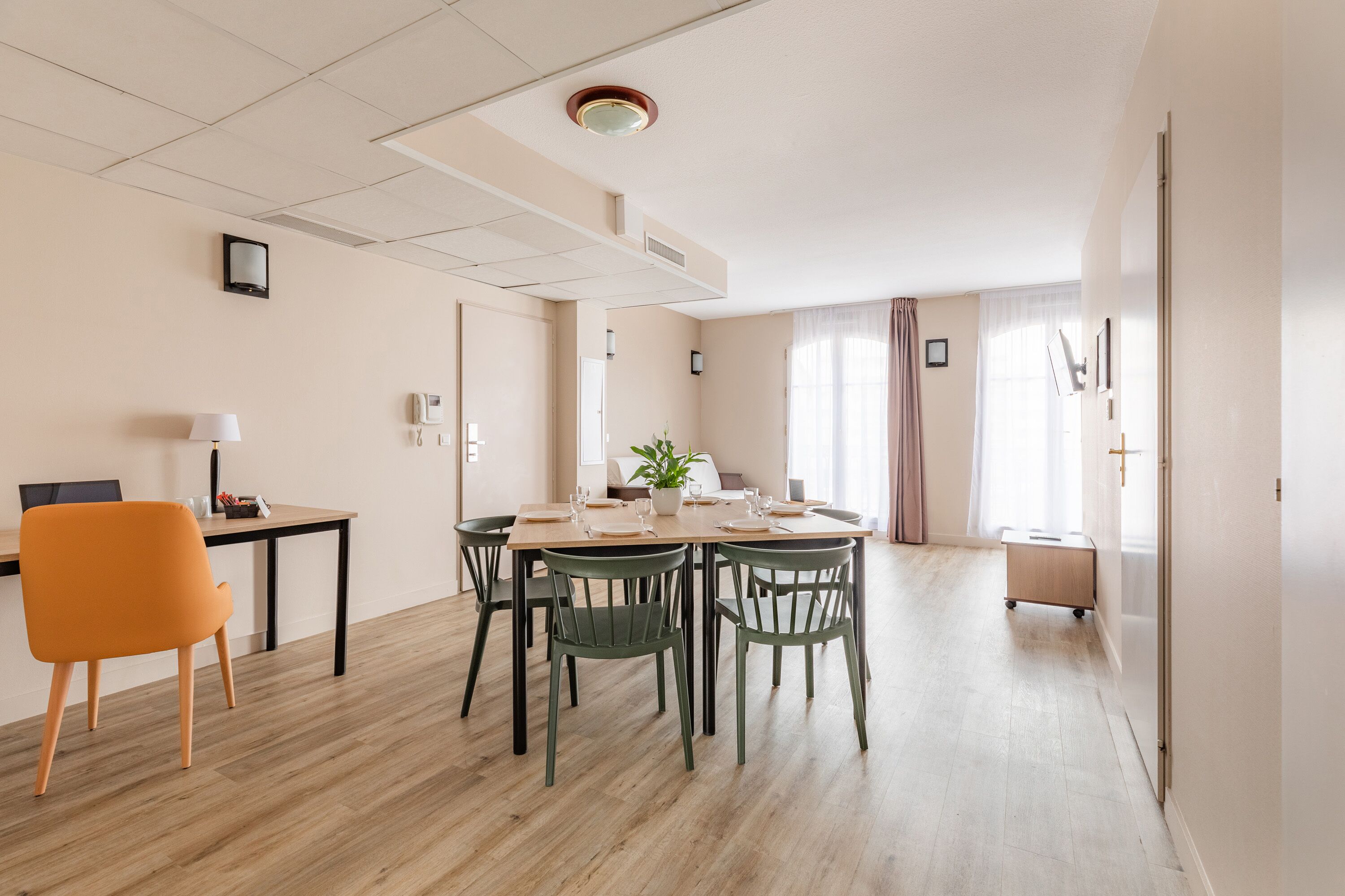 Un comedor con una mesa de madera, sillas verdes, una planta en maceta y una mesita auxiliar con una lámpara.