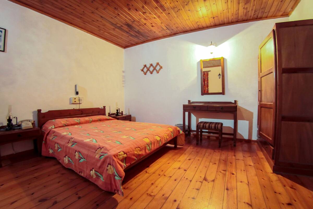 A room with a wooden floor, a bed with a floral bedspread, a desk with a chair, a mirror, and a wardrobe.