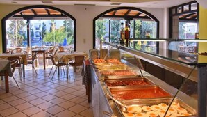 Un espace buffet proposant une variété de plats, notamment des œufs, des saucisses et des sauces, dans un espace intérieur bien éclairé, doté d’un sol carrelé et de fenêtres en arc de cercle.