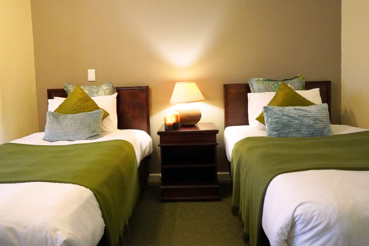 A hotel room with two beds, each with a green blanket and decorative pillows. A nightstand with a lamp and candles is between the beds.