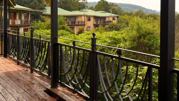 A wooden balcony with a metal railing overlooking a lush green landscape and a building with a green roof.