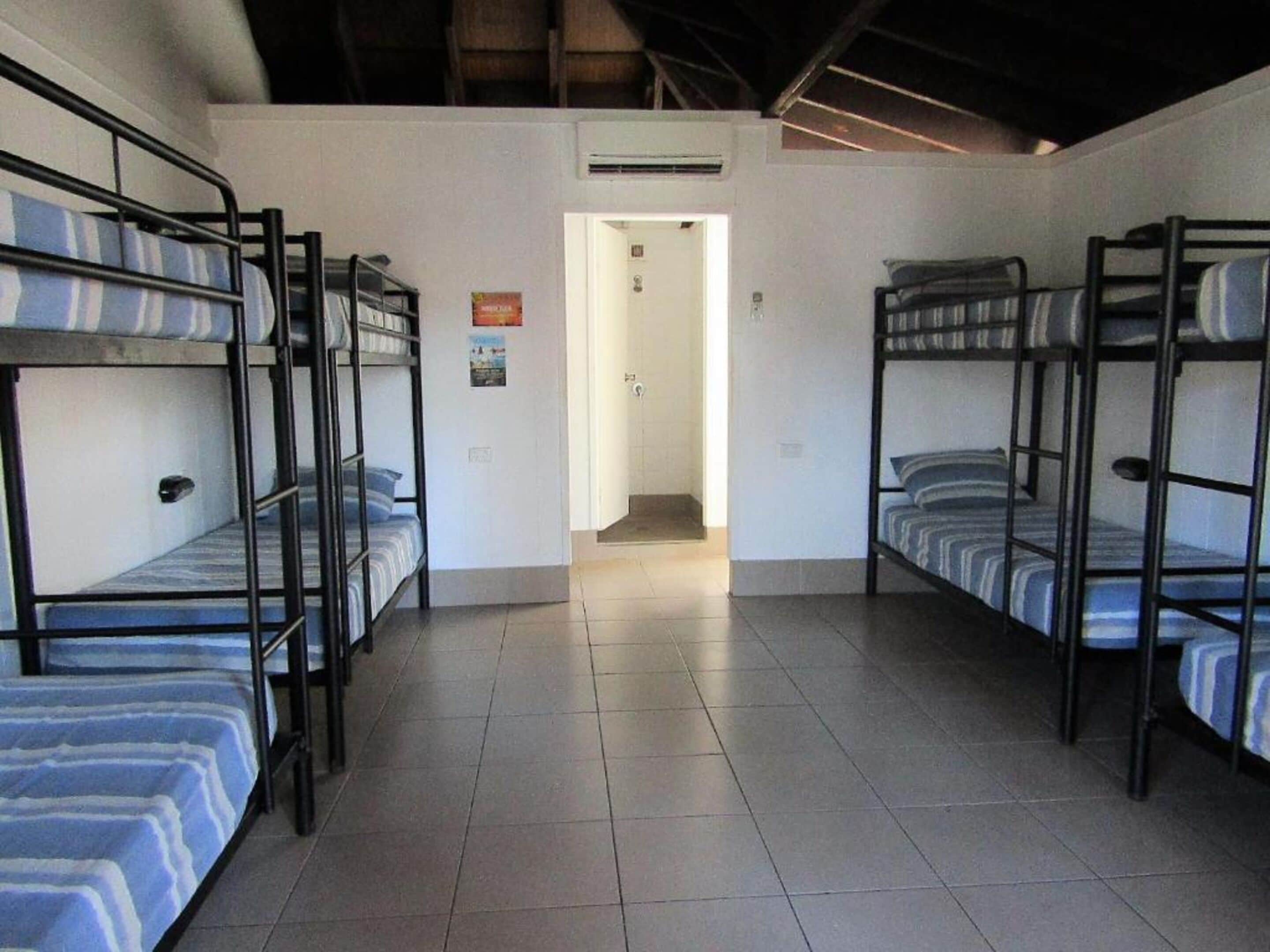 Una habitación tipo dormitorio con literas, suelo de baldosas y una puerta que da a un pasillo.