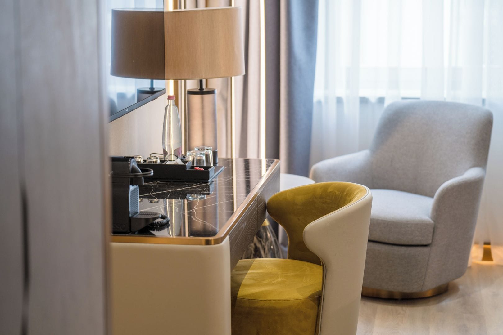 A modern hotel room with a marble-topped desk, a coffee machine, a lamp, and a comfortable armchair.