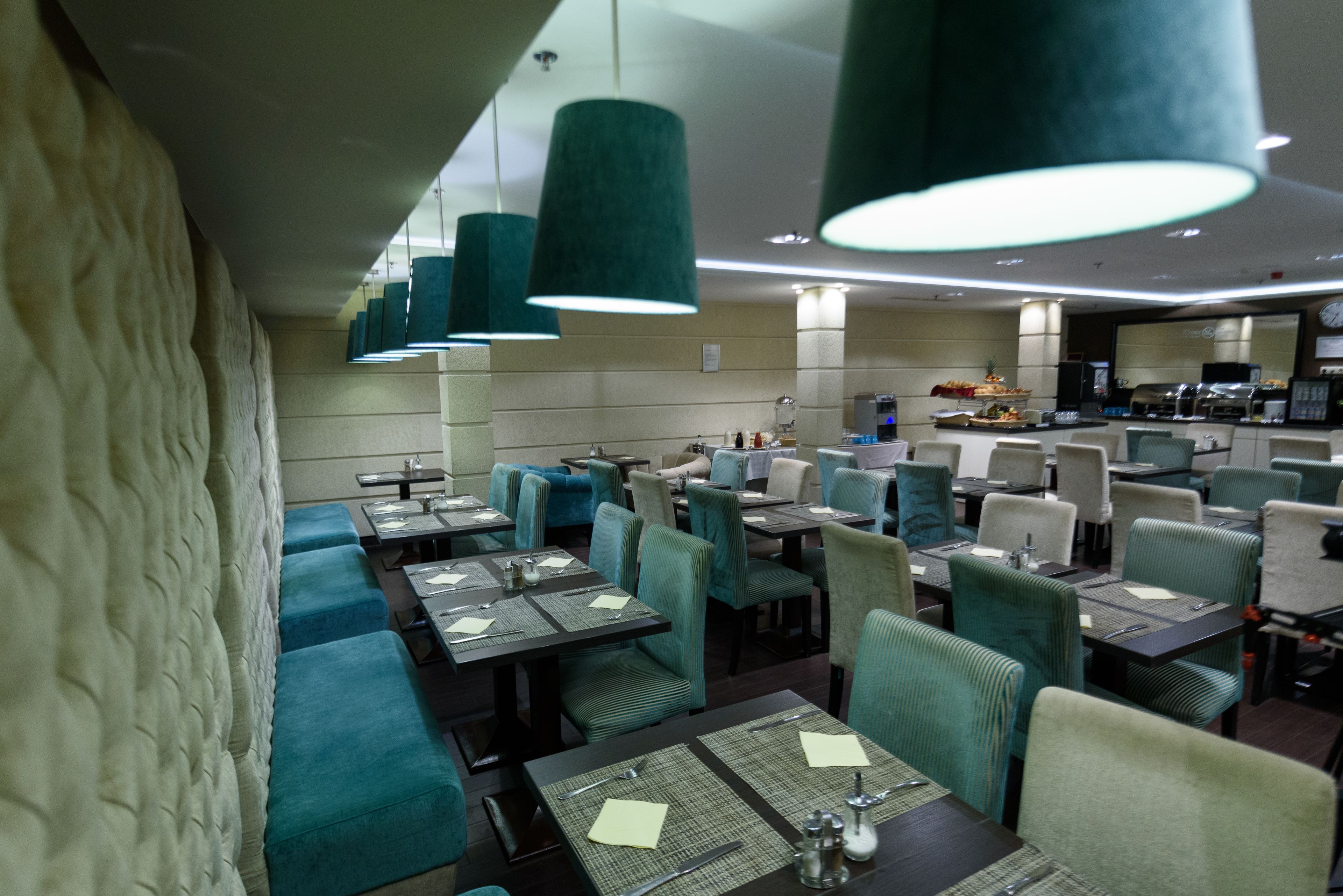 Un restaurant doté de nombreuses tables et de banquettes, de sièges recouverts d’un tissu vert et de luminaires suspendus verts.