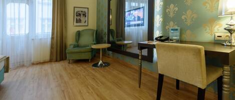 A hotel room with a green sofa, a small round table, a desk with a chair, a large mirror, and a television on the wall.