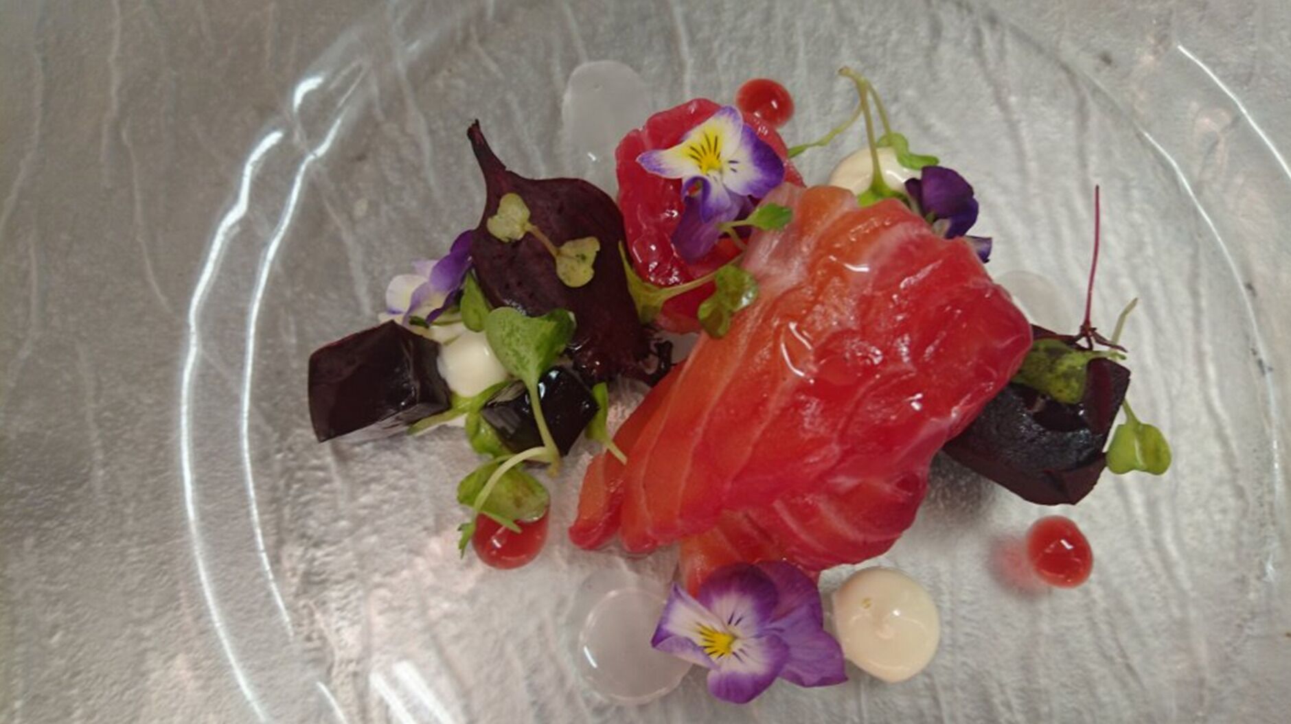 A clear glass plate with a variety of food items including salmon, greens, and flowers.