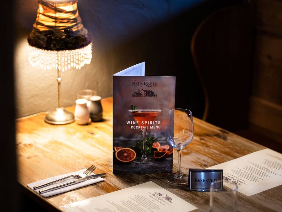 Una mesa con un menú titulado 'The BallyRobin Hotel Wine, Spirits, Cocktail Menu' y una copa de vino.