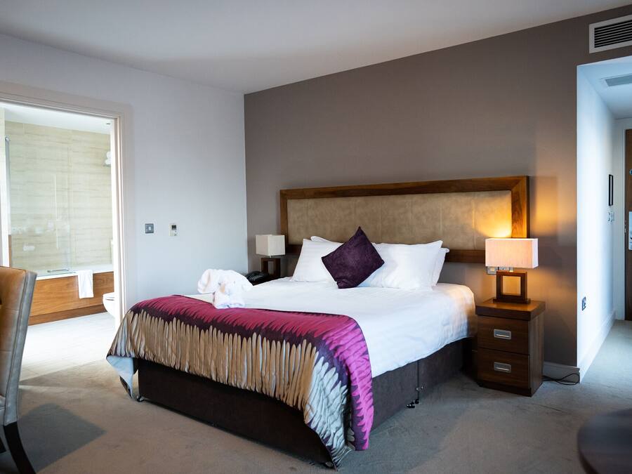 A hotel room with a large bed, a bedside table with a lamp, a chair, and a view of the bathroom.