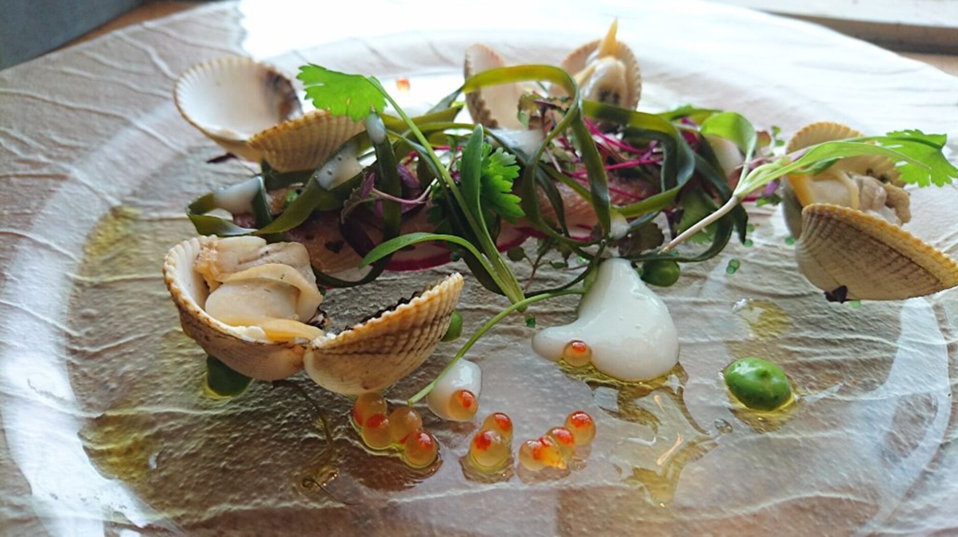 Un plato de mariscos con vieiras, verduras de hoja verde y un elemento con forma de pera sobre un plato con textura.