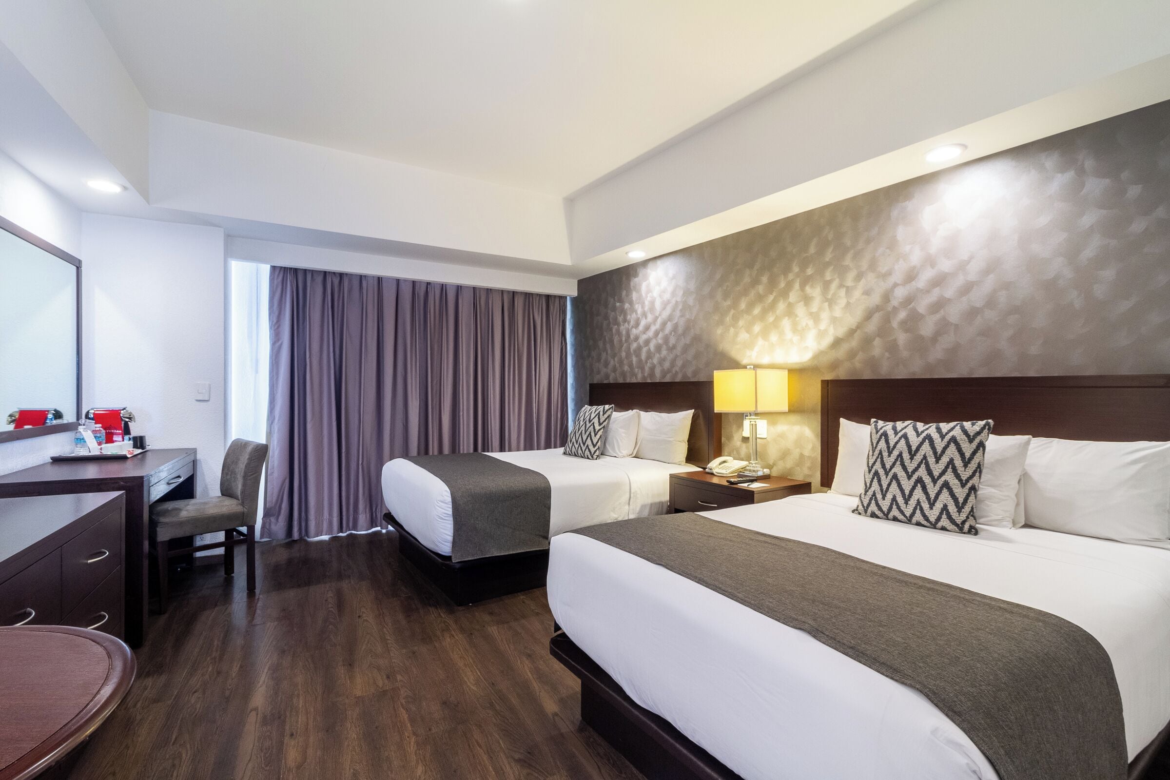 A hotel room with two beds, a desk, a chair, a desk lamp, and a window with curtains.