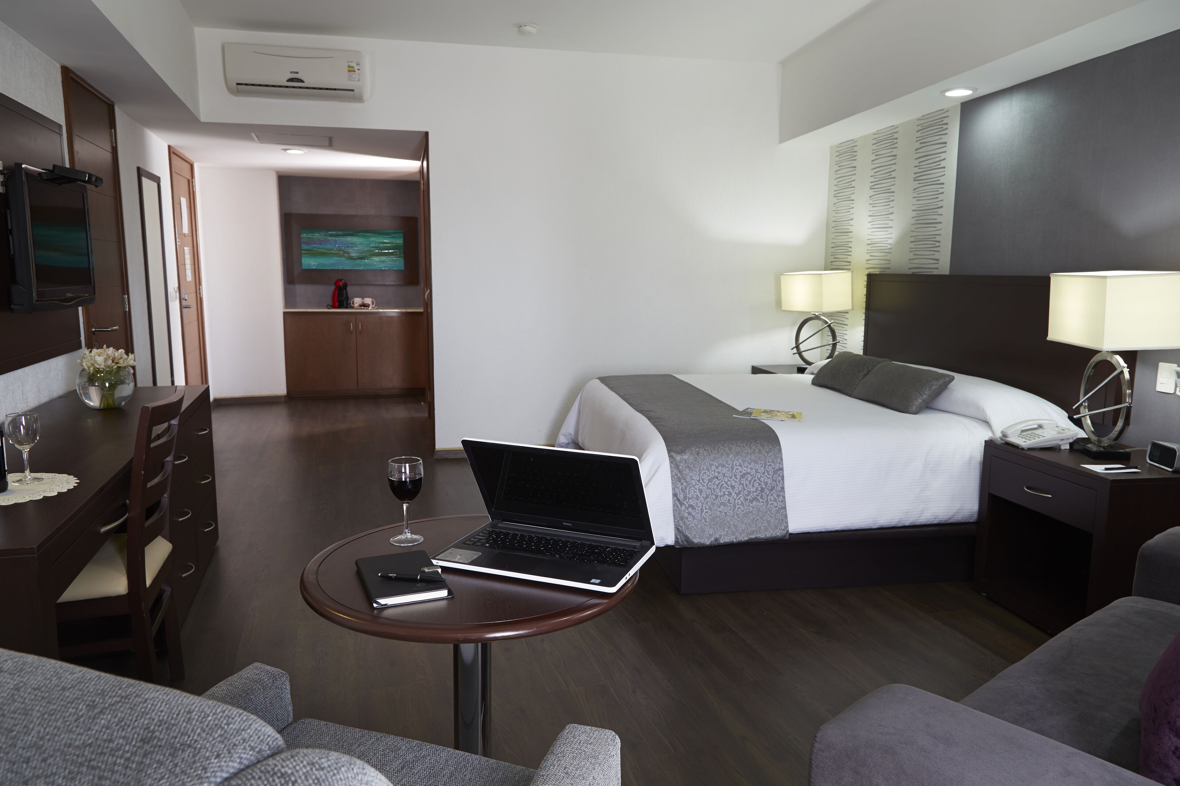 Una habitación de hotel con cama, un escritorio con una computadora portátil, una zona de comedor con mesa y sillas, y un televisor montado en la pared.