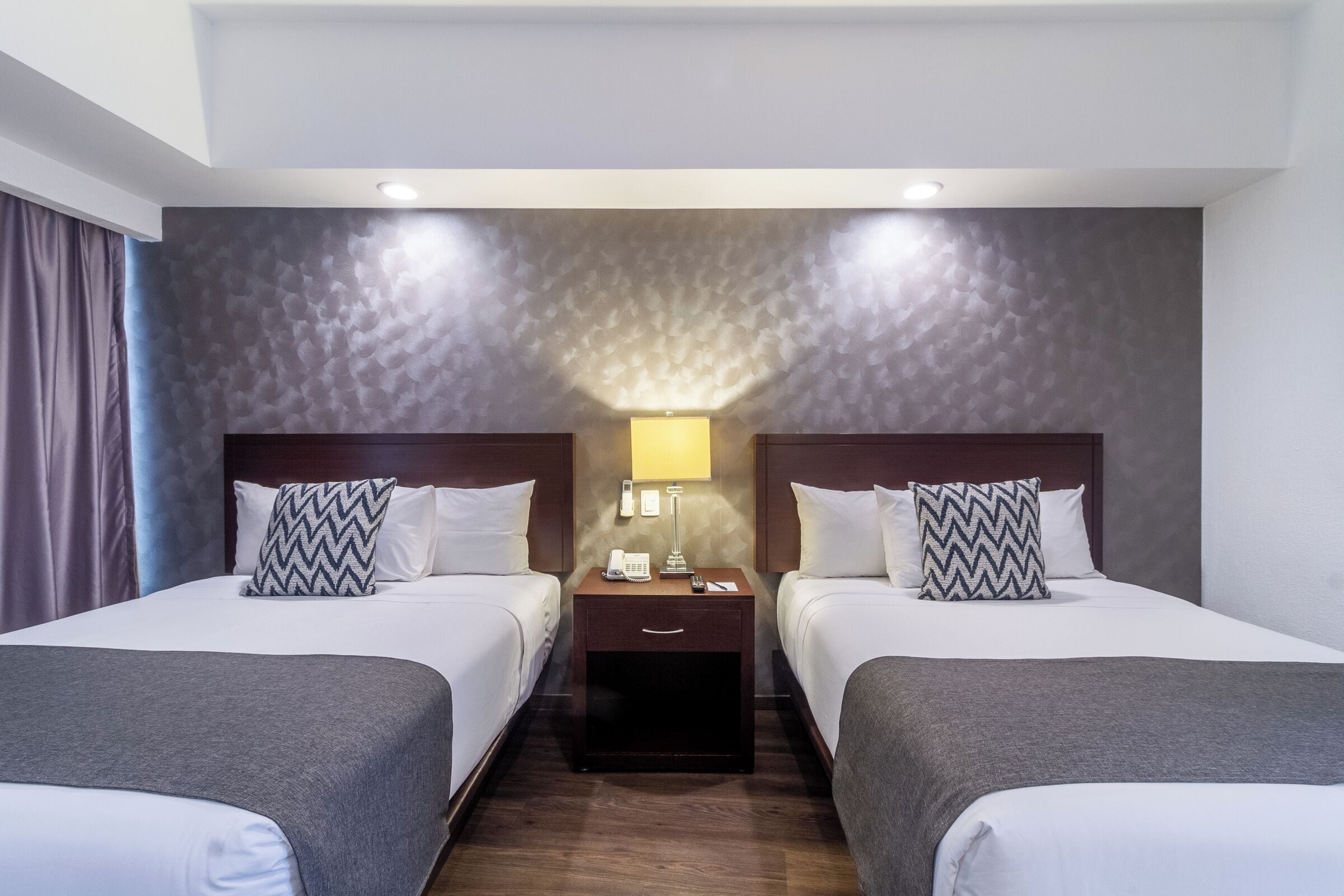 A hotel room with two beds, a nightstand with a phone and lamp, and a textured wall.