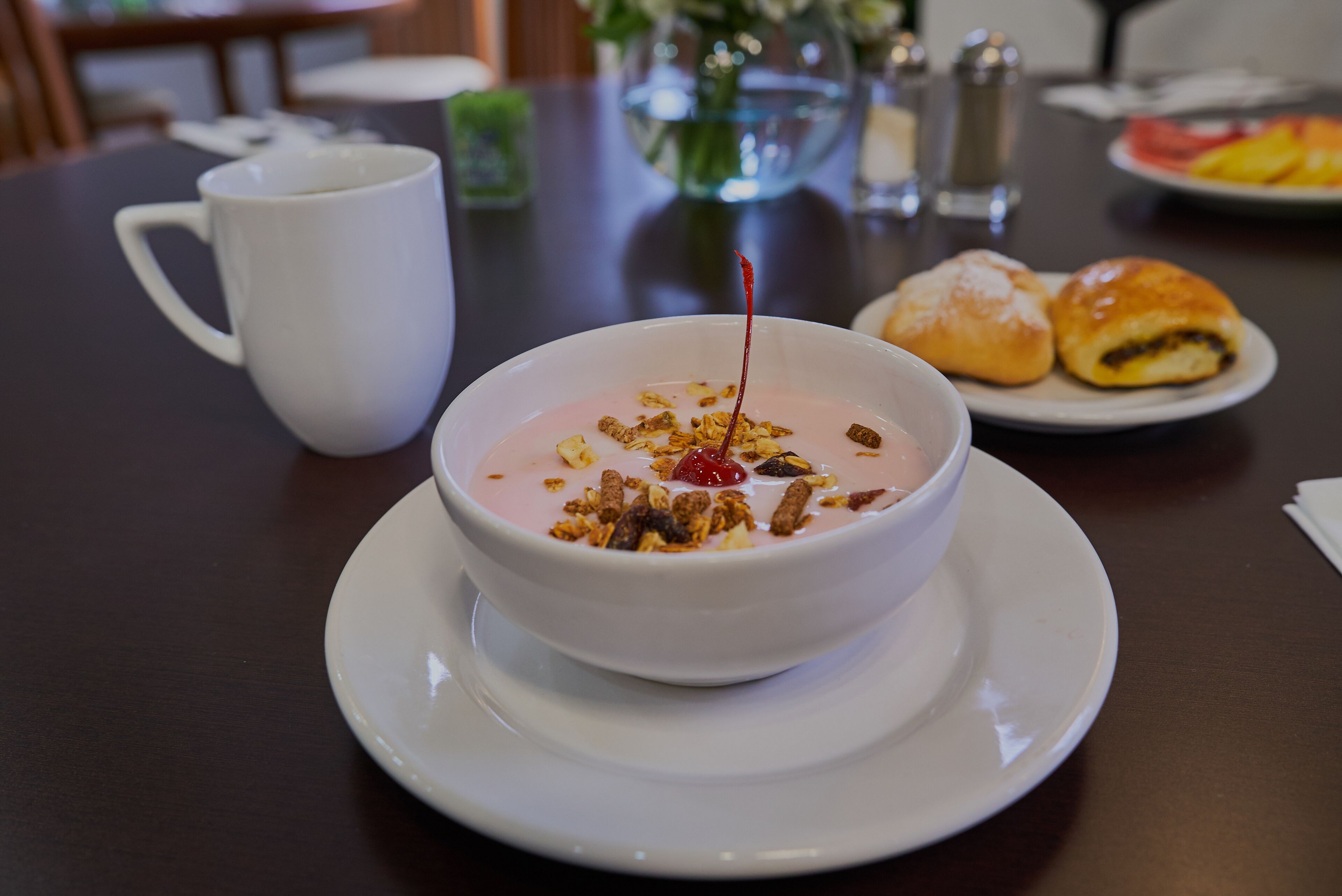 A bowl of yogurt with granola and a cherry on a white plate, a white cup, and a plate with pastries on a table.