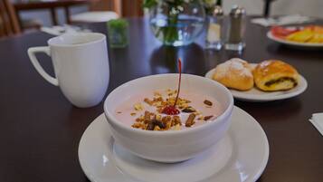 A bowl of yogurt with granola and a cherry on a white plate, a white cup, and a plate with pastries on a table.