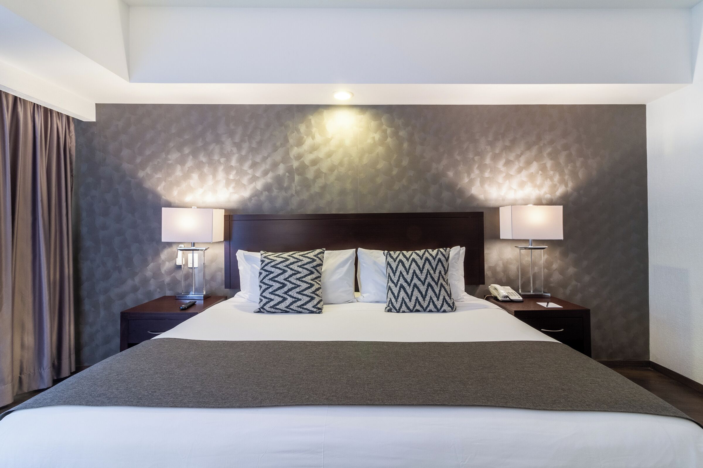 Quarto de hotel com uma cama grande, duas mesas de cabeceira e um criado-mudo com abajur e telefone.